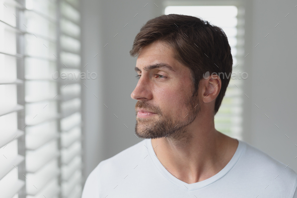 Side view of thoughtful Caucasian man looking through window blind at home