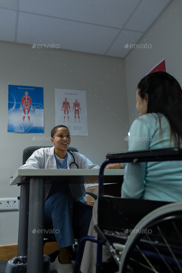Happy mixed-race female doctor talking with female patient on wheelchair at desk