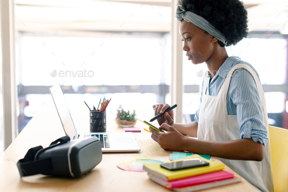 African american female graphic designer using graphics tablet while looking at laptop at desk