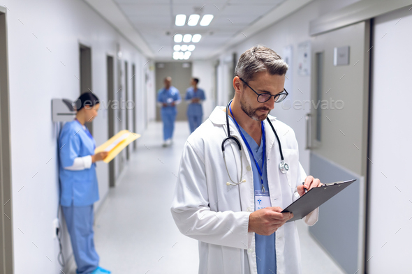 Caucasian male doctor writing on clipboard in corridor at hospital. - Stock Photo - Images