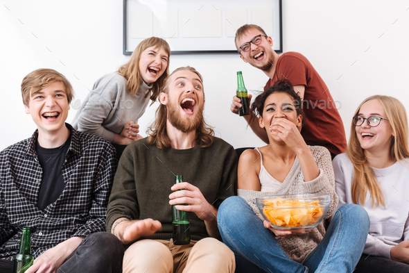 Happy laughing friends sitting on sofa and watching film together with chips and beer at home
