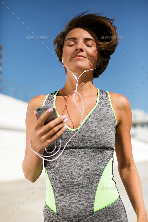 Beautiful smiling woman in modern gray sport suit jogging holding cellphone and earphones dreamily