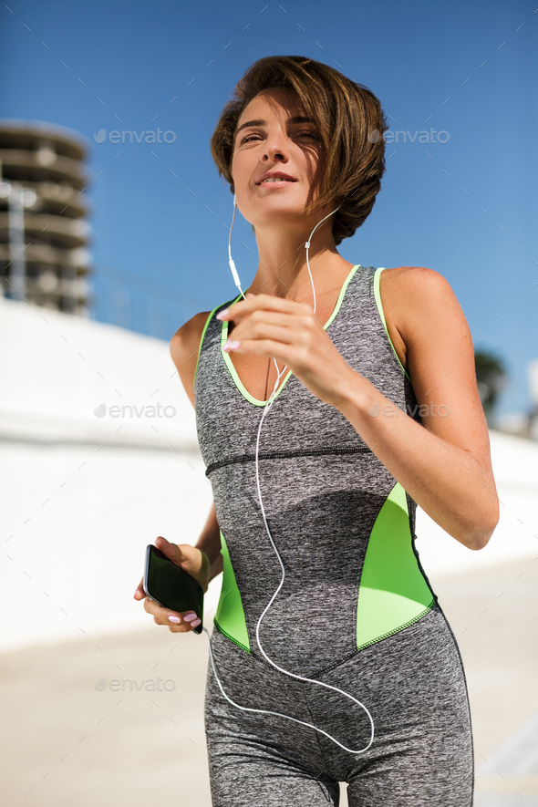 Smiling woman with short hair in modern sport suit holding cellphone and earphones while jogging