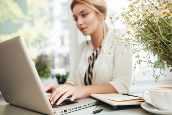 Beautiful lady with blond hair typing on computer at cafe with notebook and cup of coffee on table