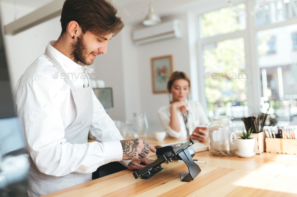 Smiling barista in apron using cash counter while girl at the counter using smartphone