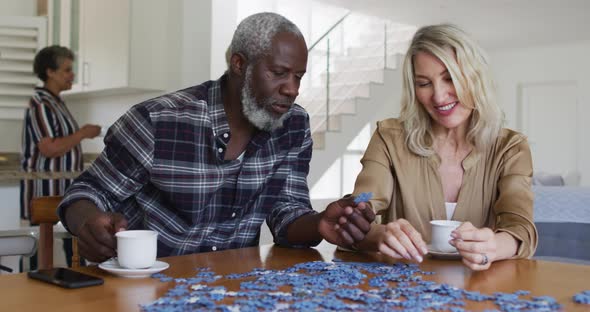 African american senior man and caucasian senior woman sitting by table doing puzzles drinking tea alt