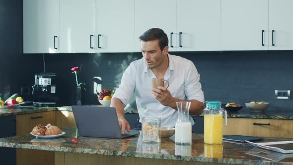 Smiling Man Watching Video Standing in Kitchen. Happy Person Scrolling Computer alt