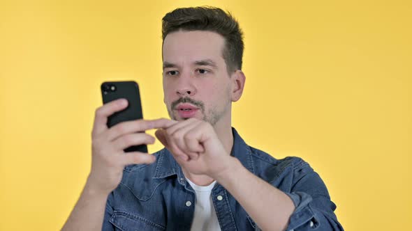 Portrait of Young Man Reacting To Loss on Smartphone, Yellow Background alt