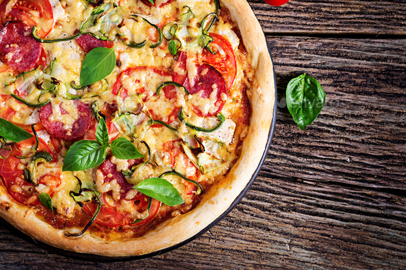 Italian pizza with chicken, salami, zucchini, tomatoes and herbs on vintage wooden background.