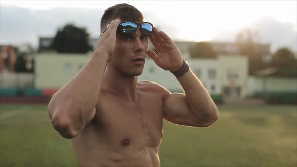 Muscular Man with a Naked Torso After Training Stands in the City Stadium and Puts on Sunglasses alt