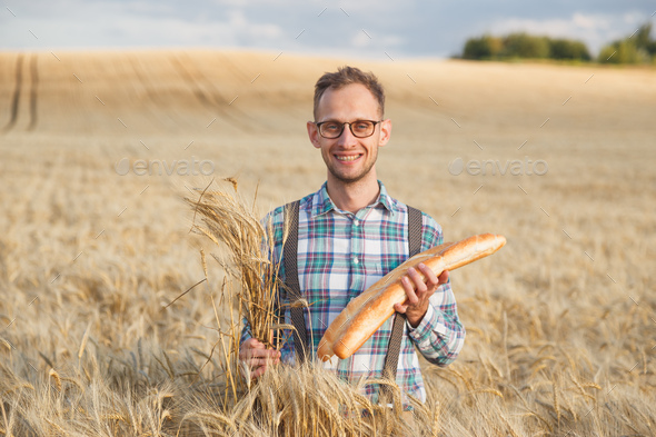 Happy farmer hold bakery products and ears of corn standing in ripe wheat field