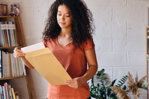 Beautiful woman in T-shirt dreamily opening envelope with exam results with bookshelf on background
