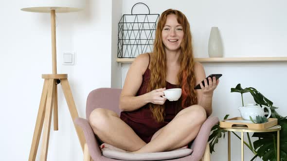 girl is sitting in room in morning on a chair with a phone drinking coffee