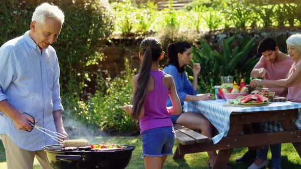 Grandfather and granddaughter preparing barbecue while family having meal alt