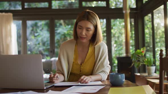 Asian woman sitting at table and working from home using laptop smiling alt