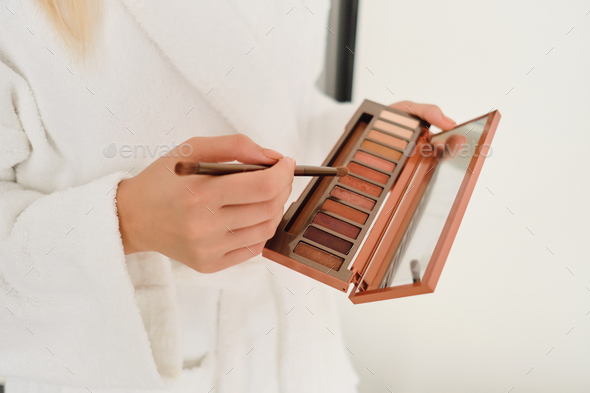 Woman in white bathrobe holding eye shadow palette with little mirror and brush in hands