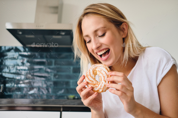 Young cheerful blond woman in white T-shirt emotionally eating sweet bun for breakfast