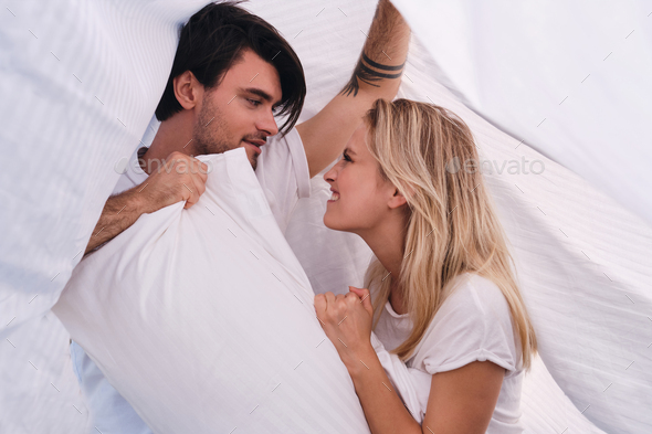 Young man and pretty emotional blond woman fighting for pillow