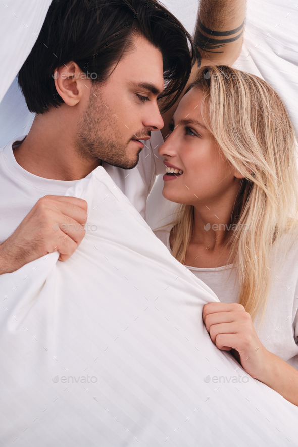 Young man and beautiful blond woman looking at each other with pillow in hands lying under blanket