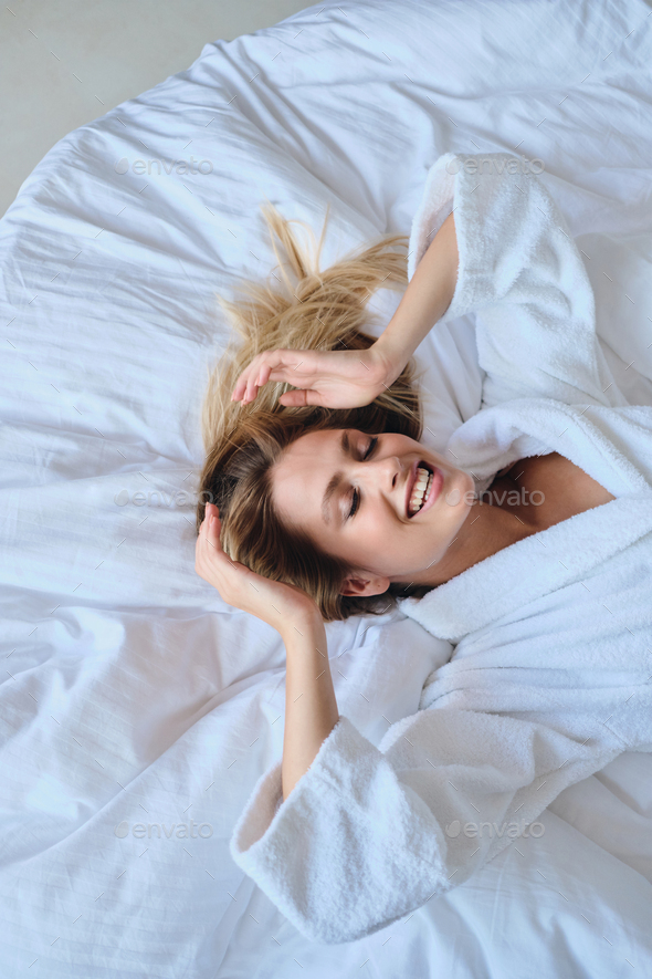 Beautiful smiling blond woman in bathrobe dreamily closing eyes lying in soft bed in hotel room