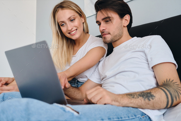 Young brunette man and beautiful smiling blond woman in white T-shirts happily using laptop together
