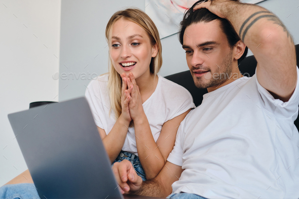 Young man and pretty smiling blond woman joyfully using laptop together sitting on bed