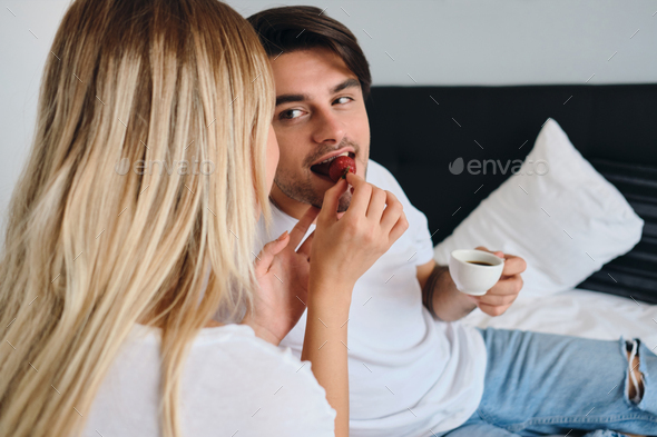 Young man holding cup of coffee in hand eating strawberry and looking at blond woman feeding him
