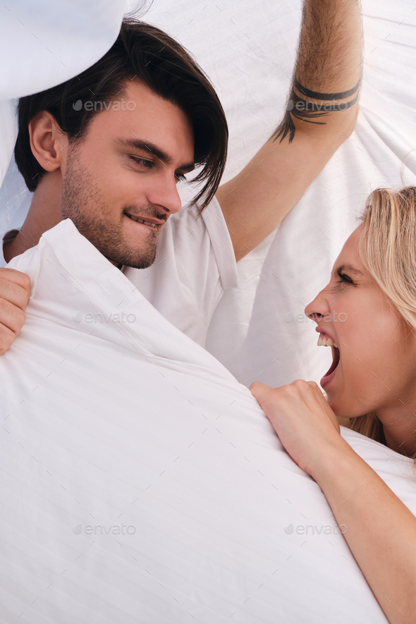 Young man and attractive emotional blond woman fighting for pillow lying under blanket