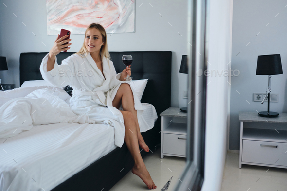 Pretty Blond Woman In Bathrobe Holding Glass Of Red Wine Happily Taking Selfie Sitting On Bed 5333