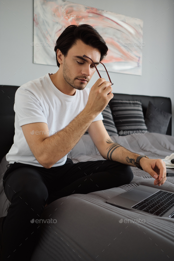 Young pensive man in T-shirt sitting on big bed thoughtfully taking off eyeglasses working on laptop