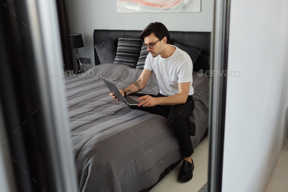 Young attractive man in T-shirt and eyeglasses in mirror reflection sitting on bed at modern home