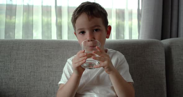Cute Baby Boy Drinking a Glass of Water Sitting on the Couch at Home alt