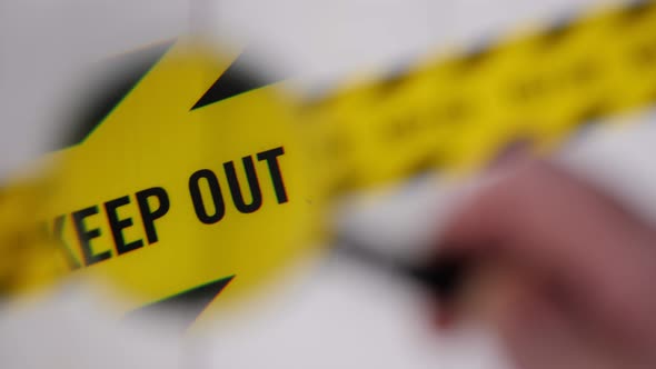 A Man Holding a Magnifying Glass Over a Yellow Barrier Tape That Says KEEP OUT alt