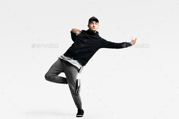 Handsome young man wearing a black sweatshirt, gray pants and a cap dancing street dances on a white