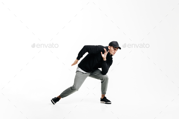 Handsome young man wearing a black sweatshirt, gray pants and a cap dancing street dances on a white