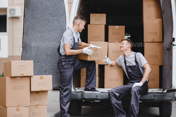 Two young handsome movers wearing uniforms are laughing while unloading the van full of boxes. House