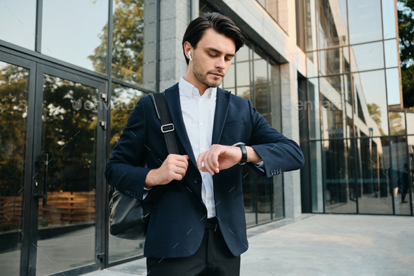 Young businessman in classic suit with earphones and backpack dreamily looking on watch outdoor