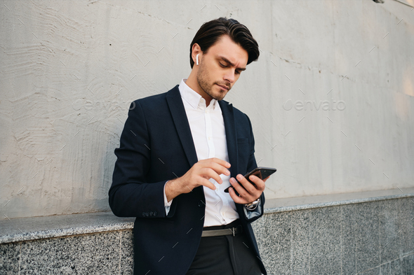 Young serious in classic suit with wireless earphones thoughtfully using cellphone outdoors - Stock Photo - Images