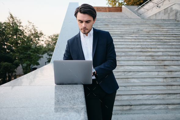 Young handsome man in classic suit standing on stairs outdoors and thoughtfully working on laptop - Stock Photo - Images