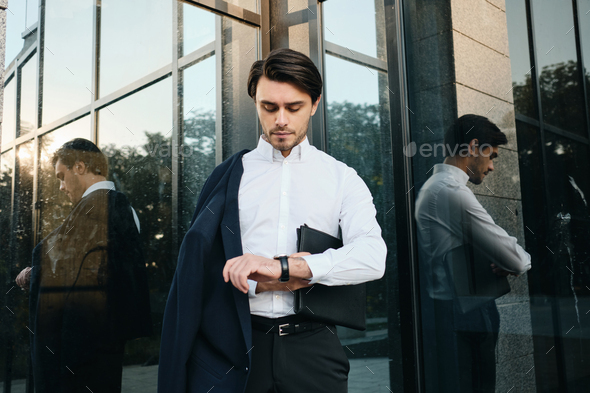 Young businessman with bag for documents thoughtfully looking on watch with over glass building - Stock Photo - Images