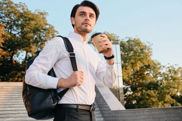 Attractive man in shirt drinking coffee carrying backpack and dreamily looking aside on city stairs - Stock Photo - Images