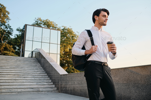 Young smiling man with backpack in white shirt drinking coffee dreamily walking in city park - Stock Photo - Images