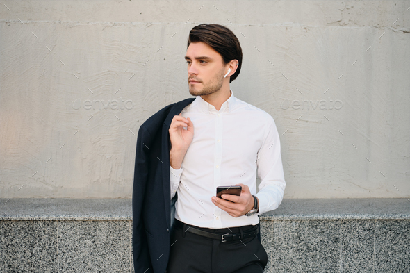 Young man in white shirt with jacket on shoulder using earphones and cellphone over gray wall - Stock Photo - Images