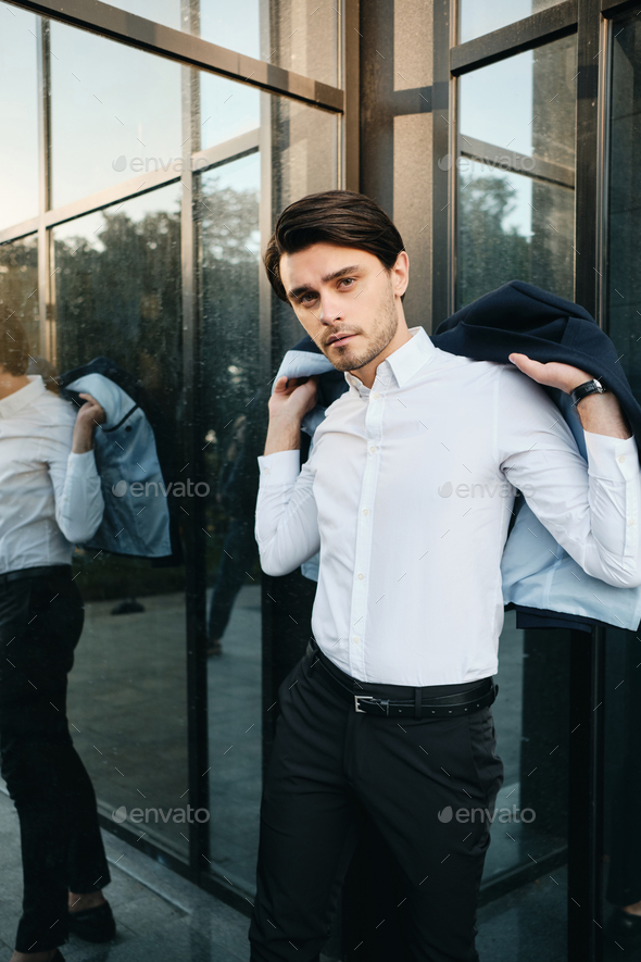 Young attractive man putting on classic jacket thoughtfully looking in camera near office centre - Stock Photo - Images