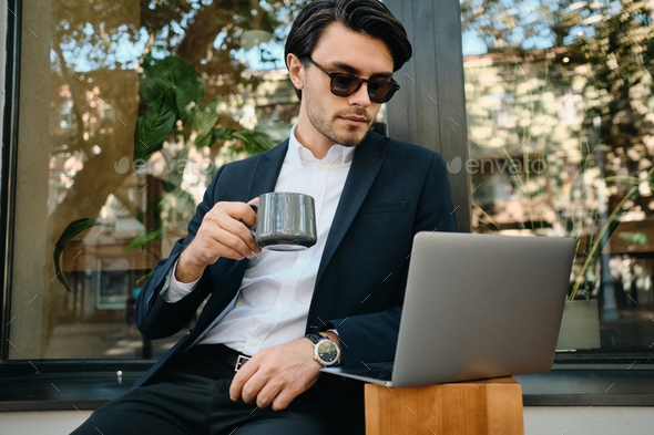 Young stylish man in jacket and sunglass drinking coffee dreamily working on laptop at street cafe - Stock Photo - Images