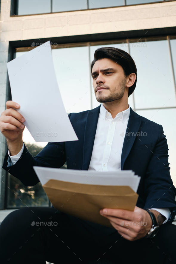 Handsome man in classic jacket sitting outdoor thoughtfully reading documents over glass building - Stock Photo - Images