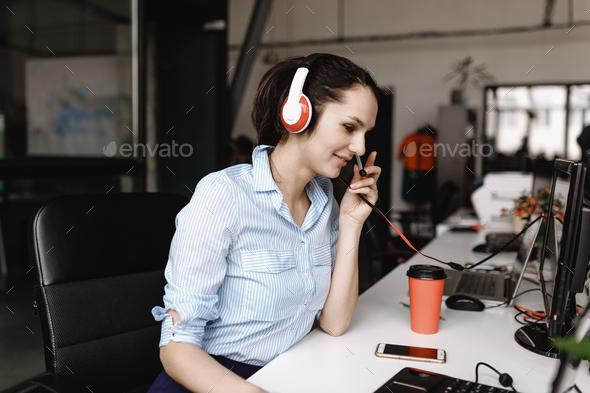 Young brunette woman dressed in office style clothes is talking to customers through the headset