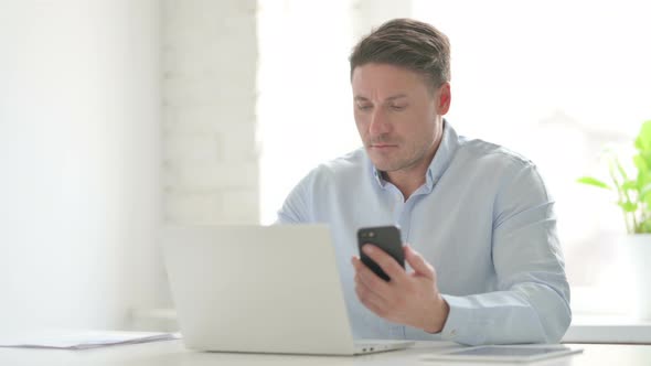 Man Browsing Internet on Smartphone while using Laptop in Office alt