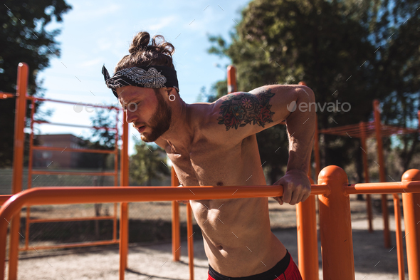 Young athletic man in headband with a naked torso with tattoos doing push-ups on the uneven bars