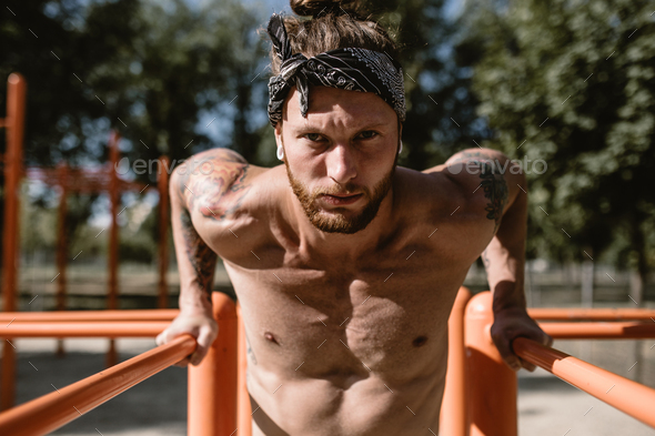 Young athletic man in headband with a naked torso with tattoos doing push-ups on the uneven bars